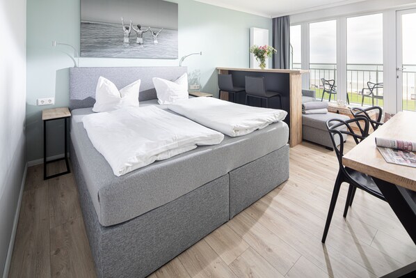 A modern hotel room with a large bed, a desk, and a balcony with outdoor seating. - Top sea view, one-room apartment Kaiserhof 312, modern, directly on the beach (Norderney)