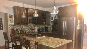 A kitchen with wooden cabinets, a granite countertop, a central island with bar stools, stainless steel appliances, and pendant lighting. - Villa Vista Mar (Sosúa)