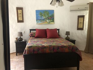 A bedroom with a bed, two nightstands, a lamp, a painting, and an air conditioning unit.