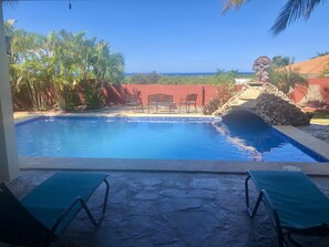 A swimming pool with two lounge chairs, a stone-tiled deck, and a view of the ocean.