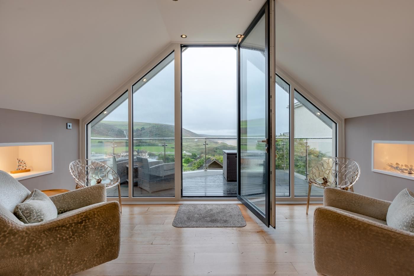 A spacious room with a large sliding glass door leading to a balcony with a view of the countryside. There are two wicker chairs and a small rug on the floor.