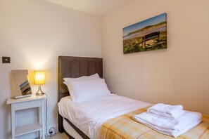 A neatly made bed with white linens and a plaid blanket, a bedside table with a lamp and a mirror, and a painting of a beach scene on the wall. - 3 Bed in Woolacombe (oc-ocvill) (Woolacombe)