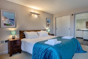 Una habitación de hotel con una cama, una mesita de noche, una lámpara de noche, una colcha azul y un cuadro en la pared.