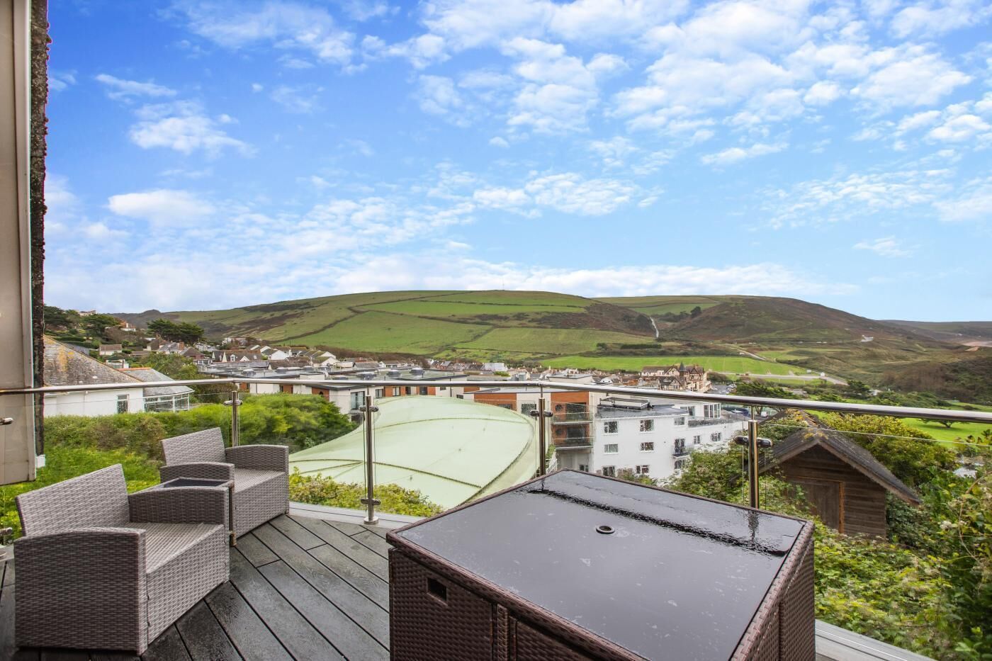 A balcony with wicker furniture overlooking a town and green hills.