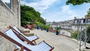 A balcony with two striped lounge chairs, a small table, and potted plants. - 4 Bed in Ilfracombe (oc-autumn) (Ilfracombe)