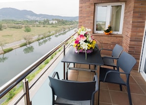 A balcony with a table and chairs, a flower arrangement, and a view of a river and distant buildings.