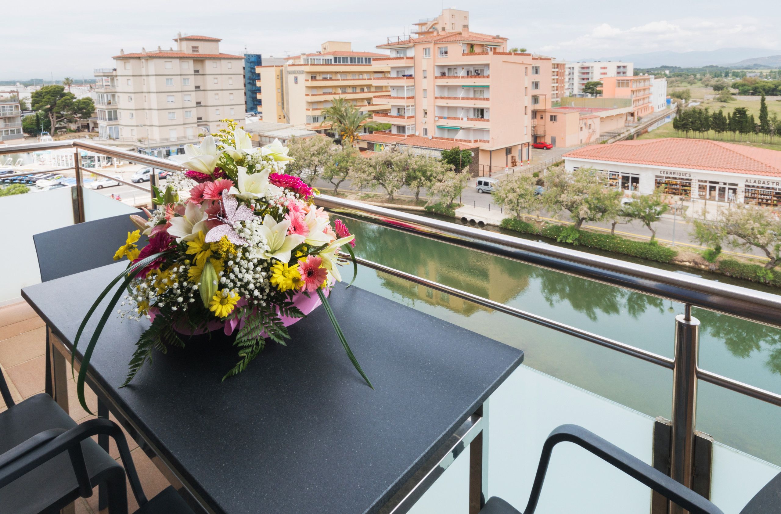 En balkon med et bord der bærer en blomsteropsætning, med udsigt over en flod og bygninger.
