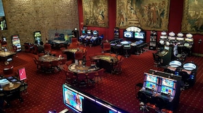 A casino with multiple gaming tables and slot machines, carpeted floors, and classical wall paintings.