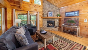 A cozy cabin interior with a stone fireplace, wooden walls, a leather sofa, and a large rug.