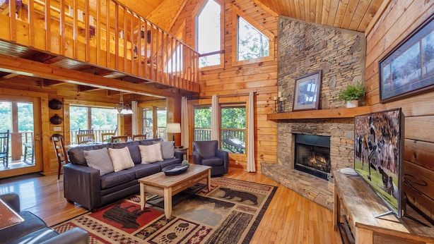 A spacious living room with a stone fireplace, wooden floors, and a large window overlooking the outdoors.