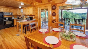 A wooden cabin interior with a kitchen, dining area, and a balcony with a view of the forest.