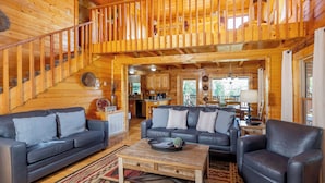 A spacious wooden cabin interior with a staircase, two sofas, a coffee table, and a dining area.