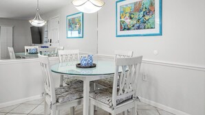 Een eethoek met een ronde glazen tafel, witte stoelen met gestreepte kussens en twee ingelijste kunstwerken aan de muur.