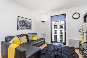 A modern living room with a black leather sofa, a black rug, a small round table, and a large window with a view of buildings. - Arco da Graça (Lisbon)
