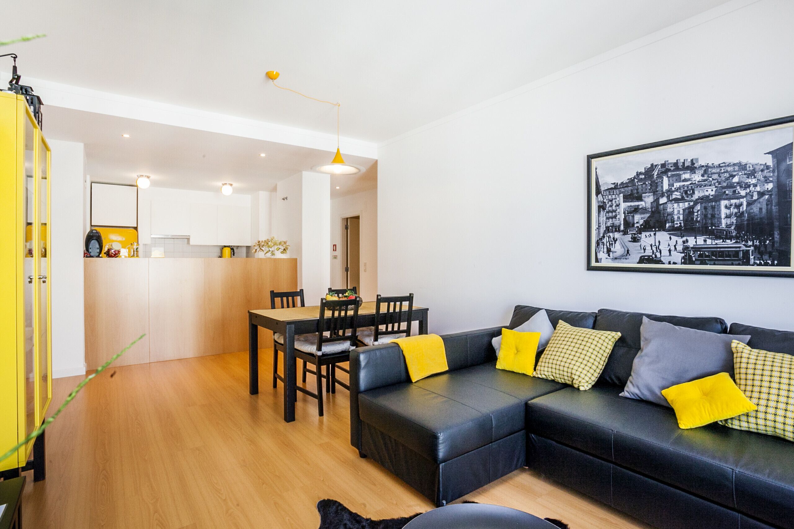 Una sala moderna con un sofá de cuero negro, cojines amarillos, una zona de comedor con una mesa y sillas de madera, y un cuadro de un paisaje urbano en blanco y negro en la pared.