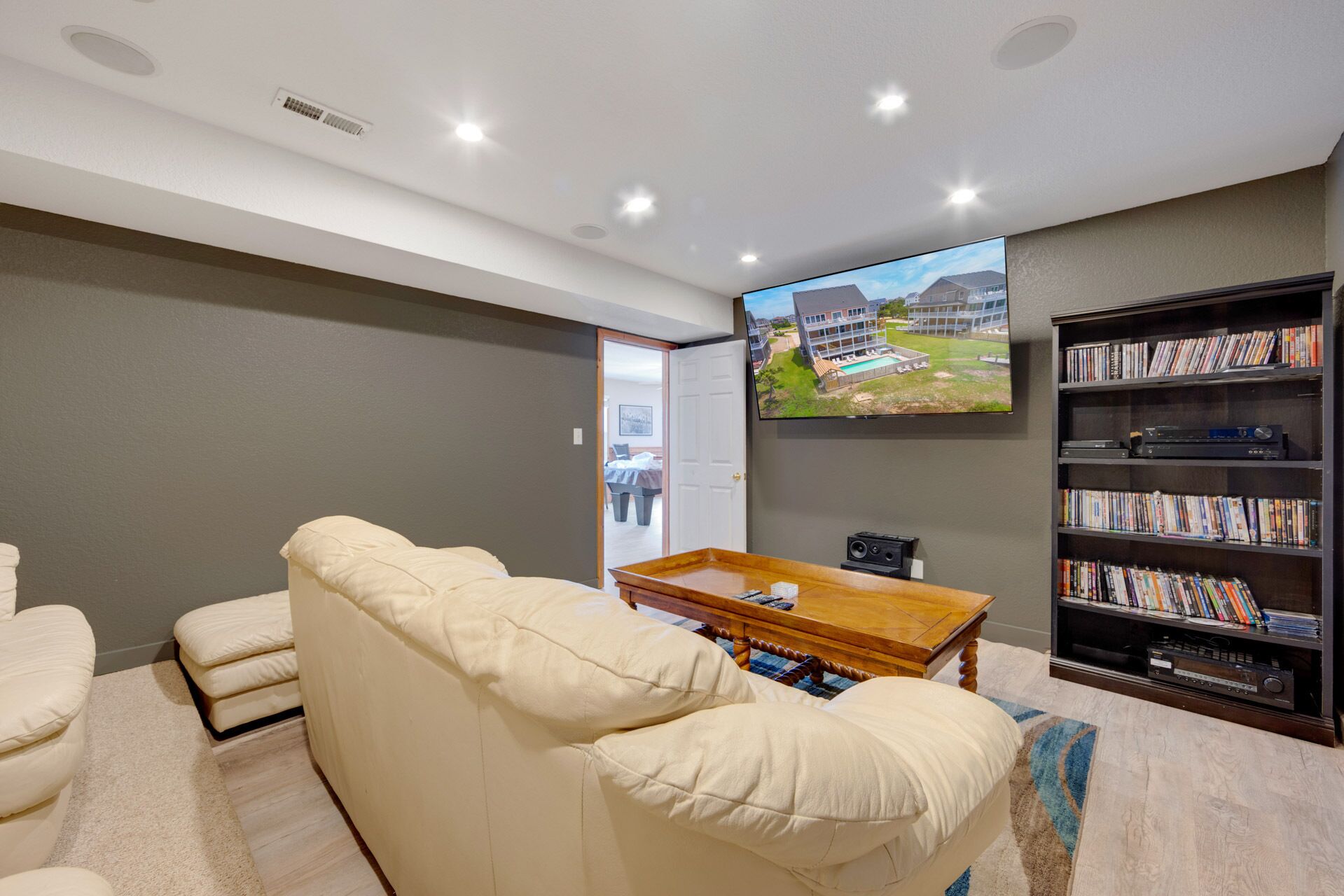 Ein Wohnzimmer mit einem beigefarbenen Sofa, einem Couchtisch, einem Bücherregal und einem Fernseher an der Wand, der einen Luftbildblick auf ein Haus zeigt.