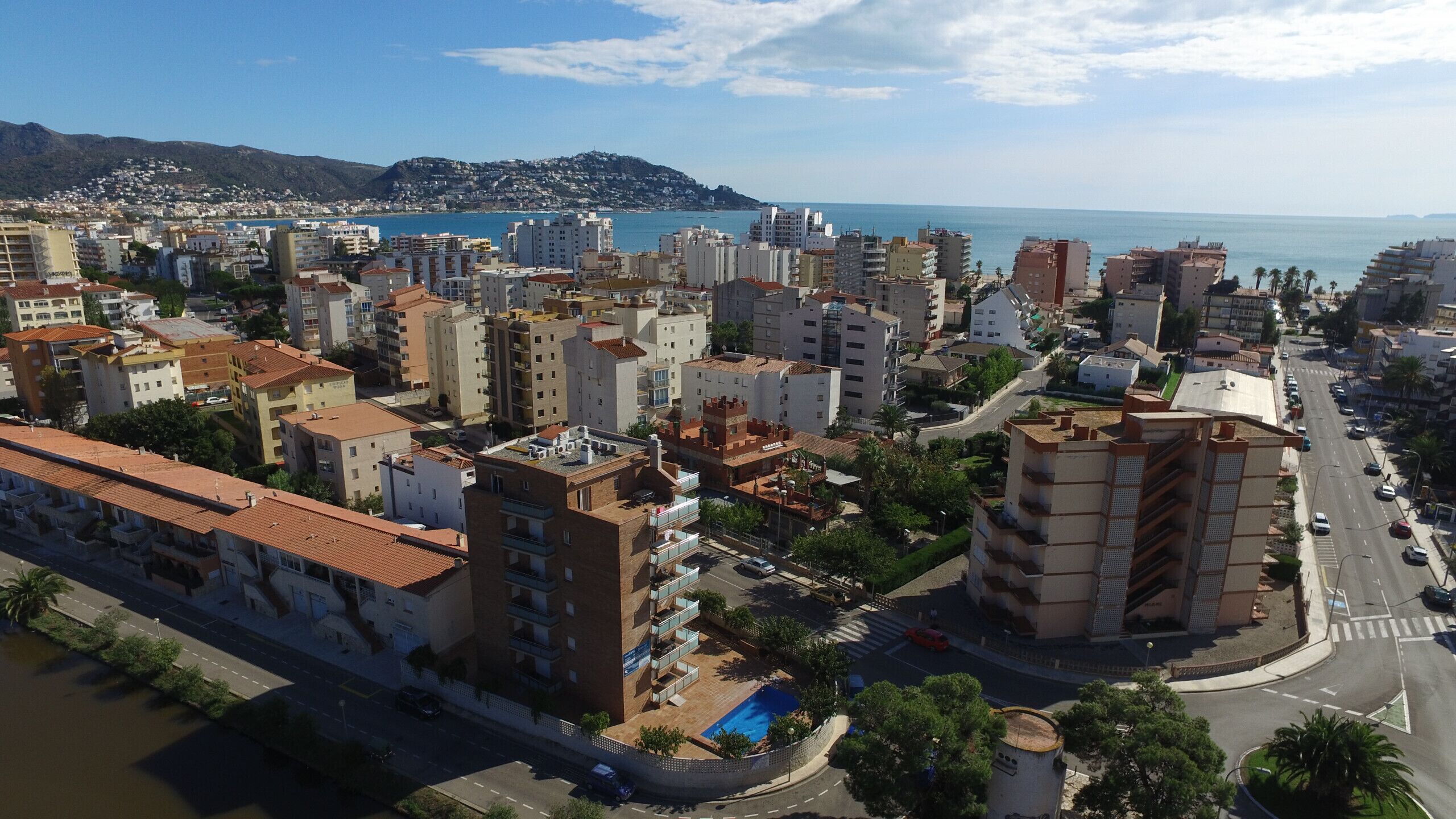 A coastal cityscape with a mix of modern and traditional buildings, a prominent pool area, and a view of the sea.