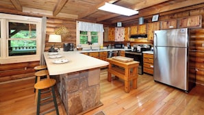 A rustic cabin-style kitchen with wooden cabinets, a central island with stools, a stainless steel refrigerator, and a large window overlooking greenery. - Very Private w/ Beautiful Scenery of Creeks! Pool, Putt Putt, Fishing, Hiking (Sevierville)