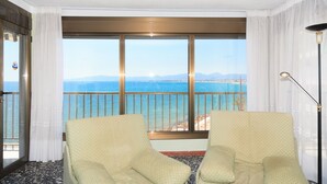 A room with a large window offering a view of the sea and distant mountains. There are two light-colored armchairs in the foreground.
