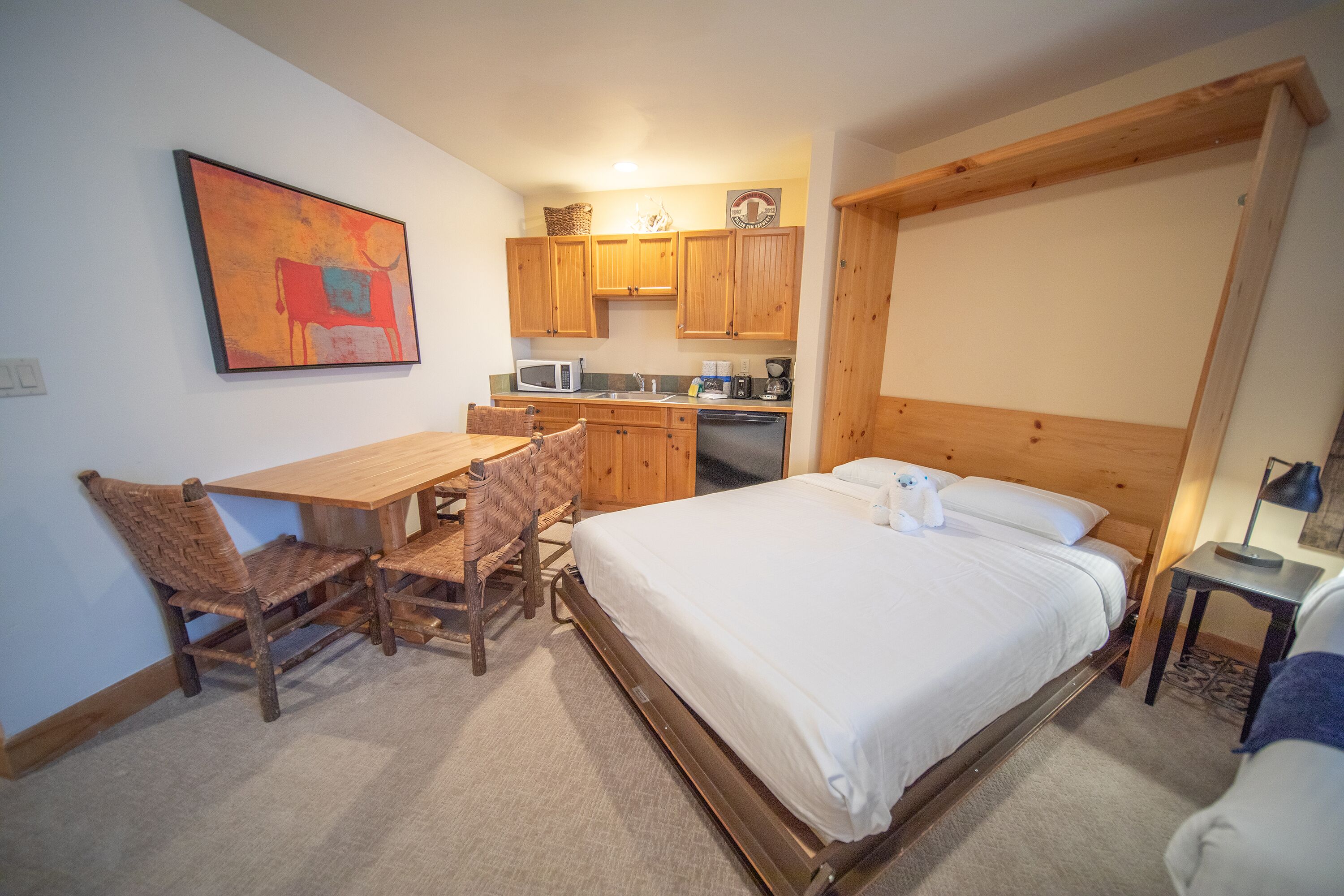 A bedroom with a wooden bed, a dining table with chairs, a kitchen area with wooden cabinets, and a painting on the wall.