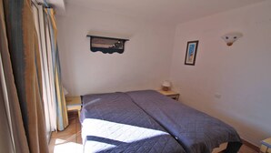 A bedroom with a bed, a nightstand, a lamp, a wall-mounted shelf with a picture, and a window with curtains.