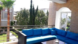 A balcony with a blue sofa and a glass table, overlooking a garden with palm trees and cypress trees.
