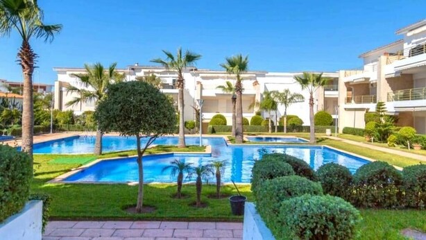 A swimming pool surrounded by palm trees and well-maintained lawns, with modern buildings in the background.