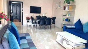 A modern living room with a sofa, a coffee table, a dining area with chairs, a TV, and a bookshelf.