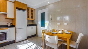 A modern kitchen with white cabinets, a built-in oven, and a refrigerator. There is a dining table with chairs and a blue door in the background. - TERRAZAS DEL DUQUE II COSTA ADEJE (Costa Adeje)