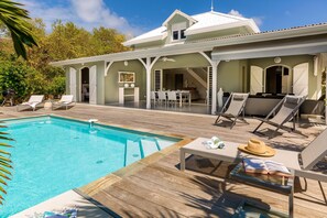 Uma área na beira da piscina com espreguiçadeiras, uma mesa com um livro e um chapéu, e vista para a casa com varanda coberta.