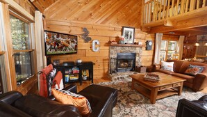 L’intérieur chaleureux d’un chalet en bois, avec une cheminée, des meubles en cuir et un téléviseur diffusant un match de football.
