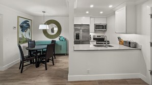 A modern kitchen with stainless steel appliances, a central island, and a dining area with a teal sofa and dark chairs. - Newly Renovated Partial Beach View 1 bed, 1 bath at Sanibel Moorings #141 (Sanibel)