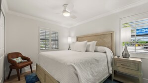 A bedroom with a bed, a chair, a nightstand, a lamp, and a window with blinds. - Newly Renovated Partial Beach View 1 bed, 1 bath at Sanibel Moorings #141 (Sanibel)
