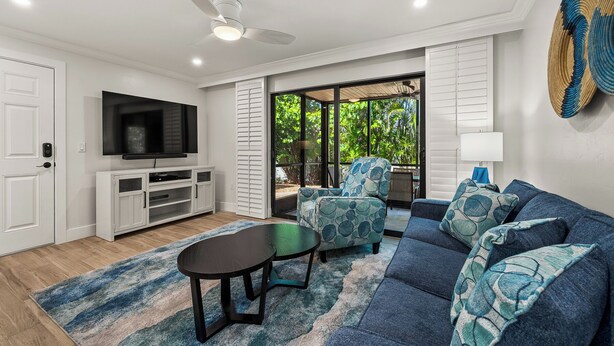 A modern living room with a flat-screen TV, a ceiling fan, a sofa set, and a view of greenery through large windows. - Newly Renovated Partial Beach View 1 bed, 1 bath at Sanibel Moorings #141 (Sanibel)