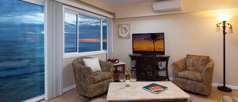 A living room with a large window overlooking the ocean, a television, and two patterned armchairs.
