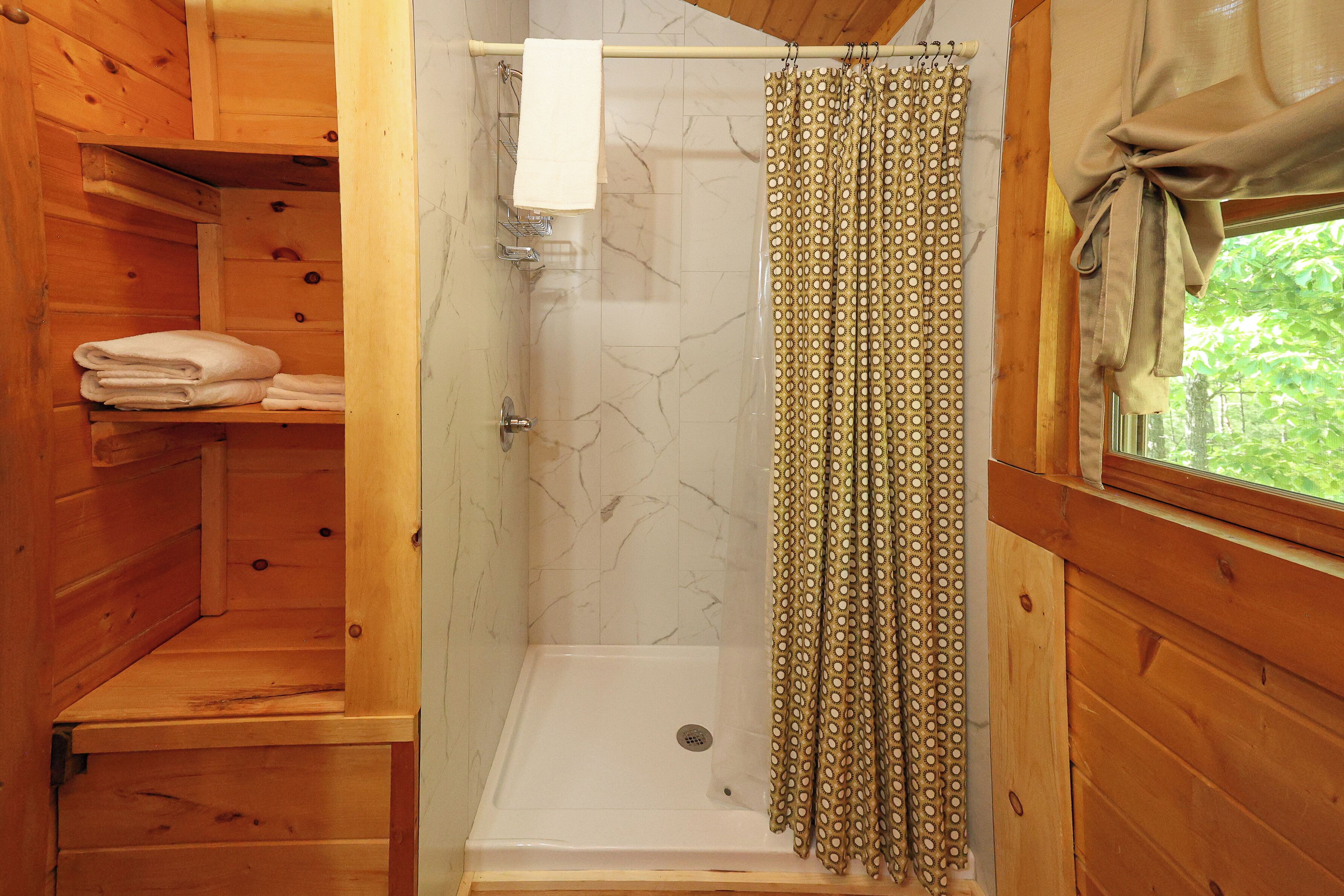 A wooden cabin bathroom with a shower, marble walls, and a window with a curtain.