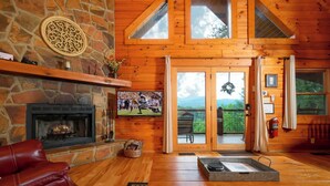 Ein gemütliches Blockhaus-Interieur mit Kamin aus Stein, Holzwänden, einem Fernseher und einem Balkon mit Blick ins Freie.
