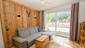 Un salon moderne avec un mur en bois, un canapé gris, une table basse en bois et une grande fenêtre donnant sur des arbres et un immeuble.