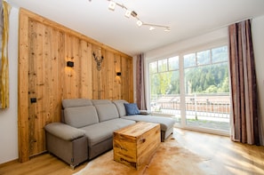 Een moderne woonkamer met een hoekbank, een houten accentmuur, grote ramen en een houten dressoir.