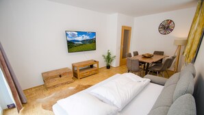 Une chambre d’hôtel moderne dotée d’un grand lit, d’un téléviseur fixé au mur, d’un coin repas avec une table et des chaises, et d’une petite armoire en bois.