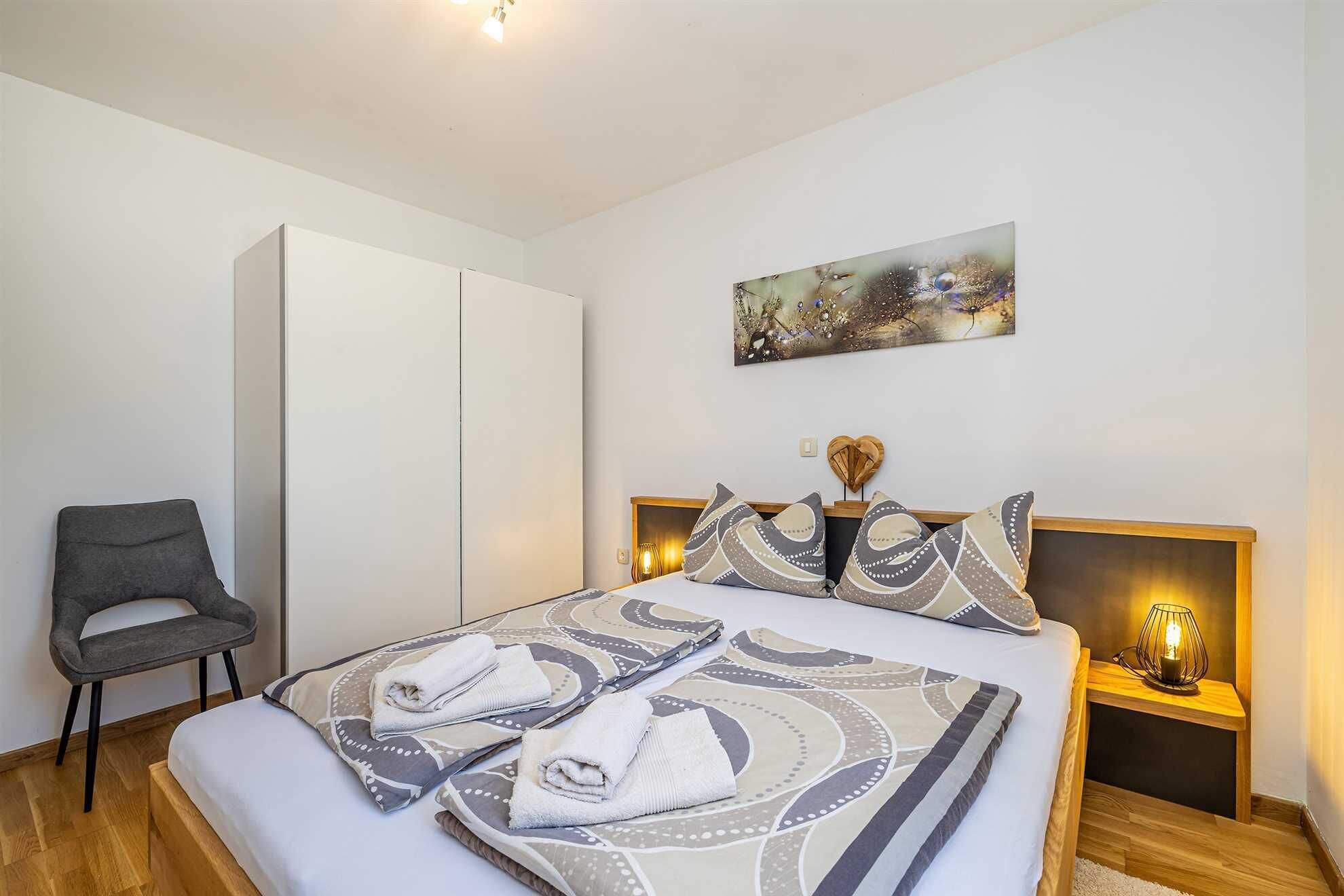 Ein Schlafzimmer mit einem Bett, einem Sessel, einem Kleiderschrank, einem Gemälde und einem Nachttisch mit Lampe.