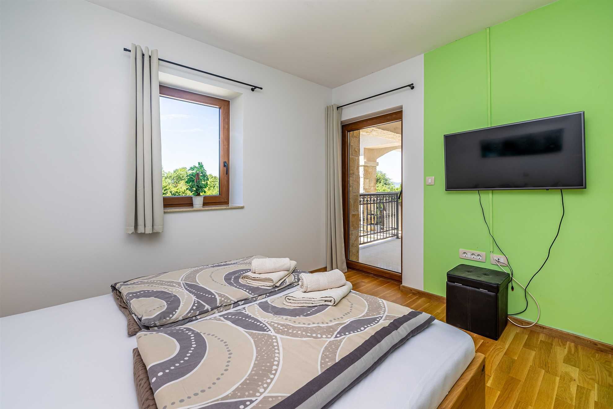 Un dormitorio con cama, televisión, ventana con cortinas y vistas al balcón.