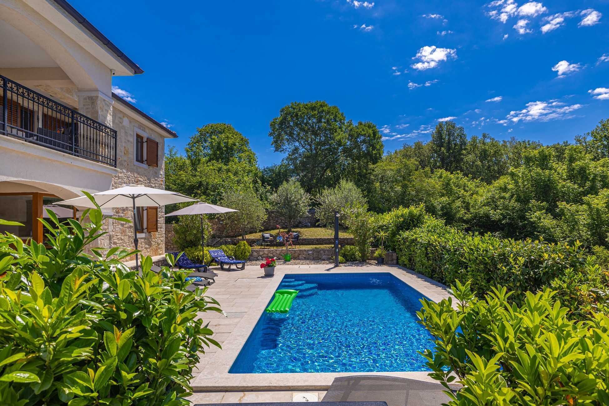 A swimming pool with lounge chairs and umbrellas, surrounded by greenery and a stone building.