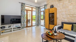 A modern living room with a flat-screen TV, a wooden cabinet with glass doors, a sofa with cushions, and a table with a fruit bowl and wine bottle. - Holiday house OBZOVA with private heated pool (Ljutići)