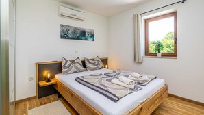 A bedroom with a wooden bed, a nightstand, a wall-mounted air conditioner, a window with a view of greenery, and a painting on the wall. - Holiday house OBZOVA with private heated pool (Ljutići)