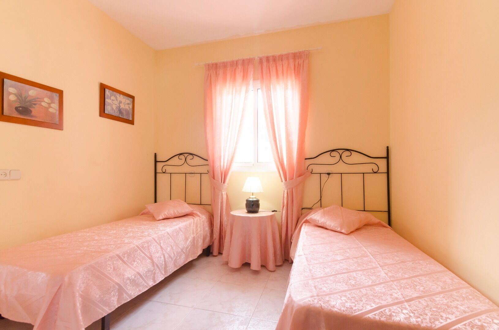 Une chambre avec deux lits simples, une petite table avec une lampe et une fenêtre avec des rideaux roses.