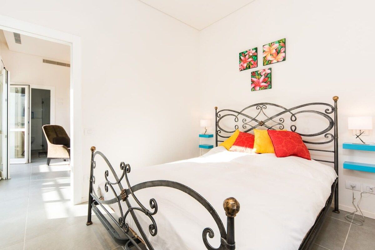 Un dormitorio con estructura de cama metálica, ropa de cama blanca y una silla en la esquina.