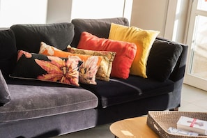 A dark-colored sofa with multiple cushions in various colors and patterns, a small wooden side table with a tray and a book, and a glimpse of a window in the background.