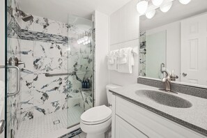 Une salle de bain avec une douche à parois de verre, des toilettes blanches, un lavabo sur un plan de travail en granit et un grand miroir.