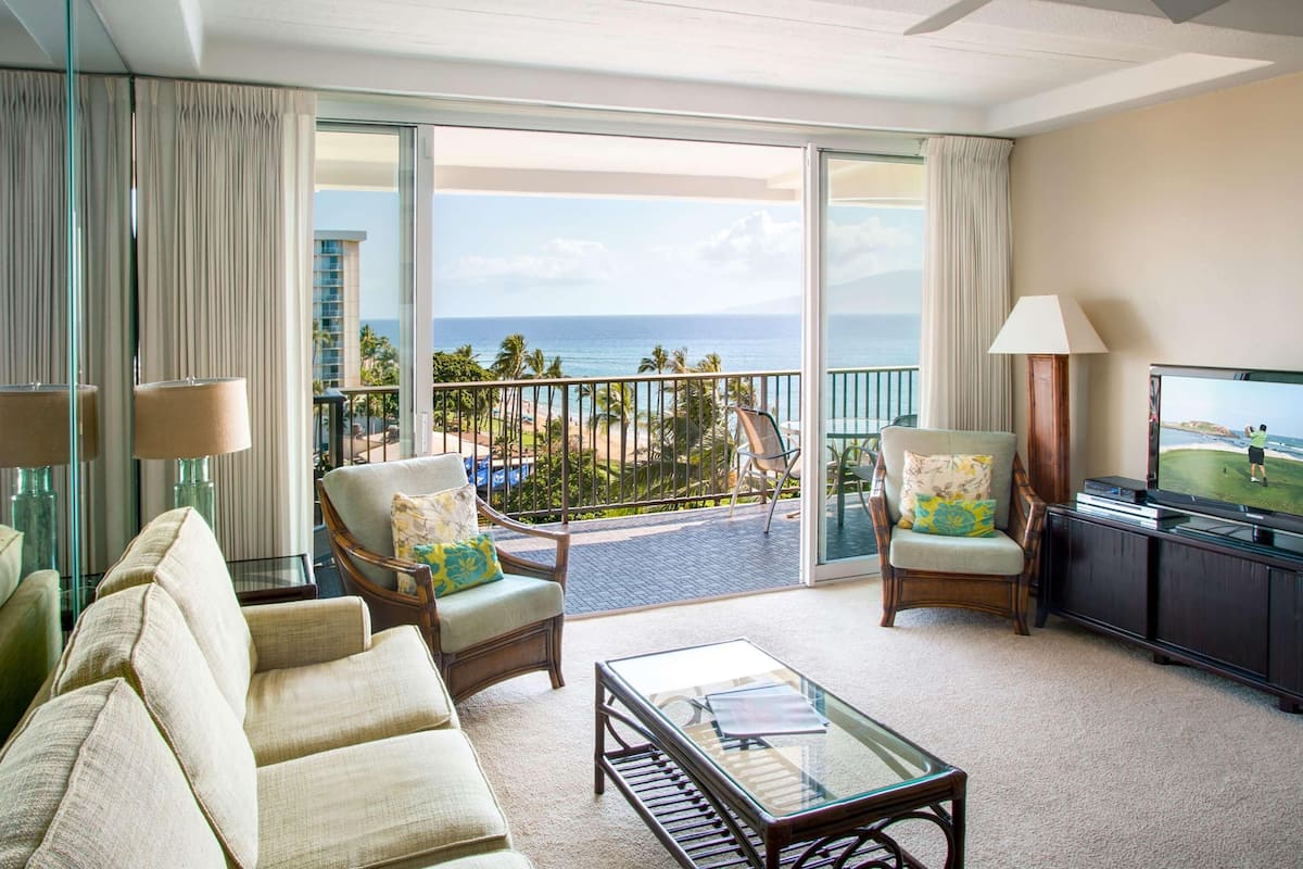 Ein Hotelzimmer mit Balkon, zwei Sesseln, einem Sofa, einem Couchtisch, einem Fernseher und Blick auf den Ozean.